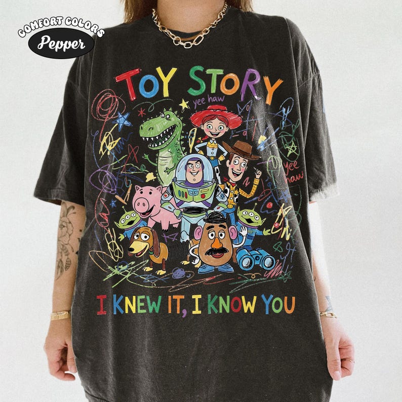 Puede incluir: Una camiseta gris carb&oacute;n de manga corta con un estampado gr&aacute;fico colorido de personajes de Toy Story, incluyendo a Woody, Buzz Lightyear, Jessie, Rex, Hamm, Slinky Dog, Mr. Potato Head y alien&iacute;genas, con l&iacute;neas garabateadas y el texto TOY STORY.