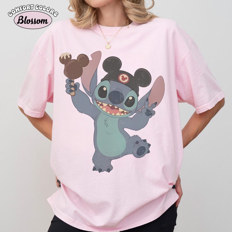 May include: A light pink short-sleeve t-shirt featuring a graphic of the character Stitch wearing black mouse ears and holding a brown chocolate-covered treat. The text Comfort Colors Blossom is printed in the upper left corner of the image.
