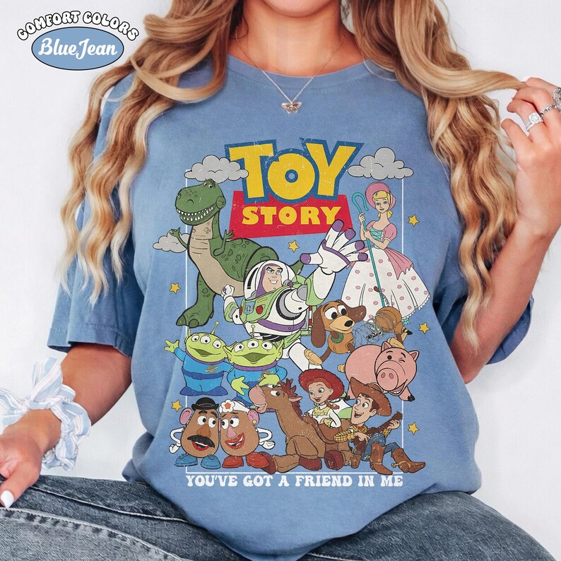 May include: A blue short-sleeve t-shirt featuring a graphic print of Toy Story characters including Buzz Lightyear, Woody, Rex, and Bo Peep. The design includes the text TOY STORY and YOU'VE GOT A FRIEND IN ME. The shirt has a crew neckline.