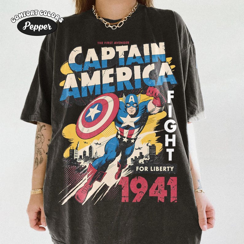 May include: A charcoal gray t-shirt featuring a graphic print of Captain America in a dynamic pose with a shield. The text reads THE FIRST AVENGER, CAPTAIN AMERICA, FIGHT FOR LIBERTY, and 1941. A Comfort Colors Pepper label is visible in the top corner.
