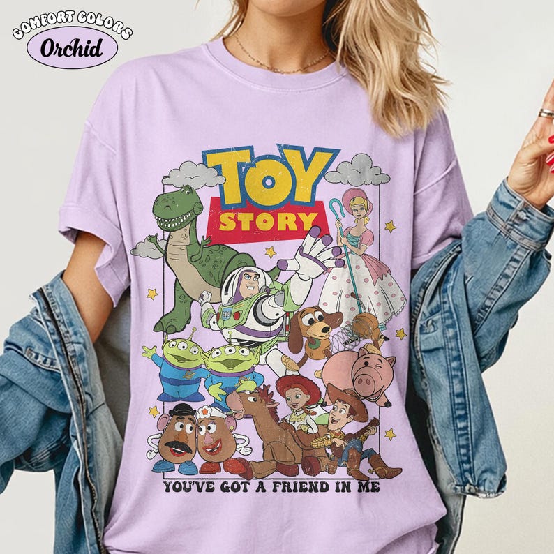 May include: A light purple t-shirt featuring a graphic print of Toy Story characters including Buzz Lightyear, Woody, Rex, Bo Peep, and others. The text reads TOY STORY and YOU'VE GOT A FRIEND IN ME. The shirt has a crew neckline and short sleeves.