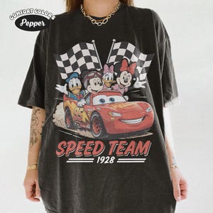 May include: Black Comfort Colors t-shirt with a Disney-themed race car graphic. The design includes Mickey Mouse, Donald Duck, Daisy Duck, and Minnie Mouse, with checkered flags. The text "SPEED TEAM 1928" is printed below the car.