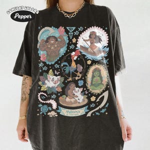 May include: Black Comfort Colors t-shirt featuring a colorful graphic of characters from the animated film "Moana." The design includes images of Maui, Moana, Pua, Hei Hei, and other elements from the movie. The text "Moana & Friends" is also visible.