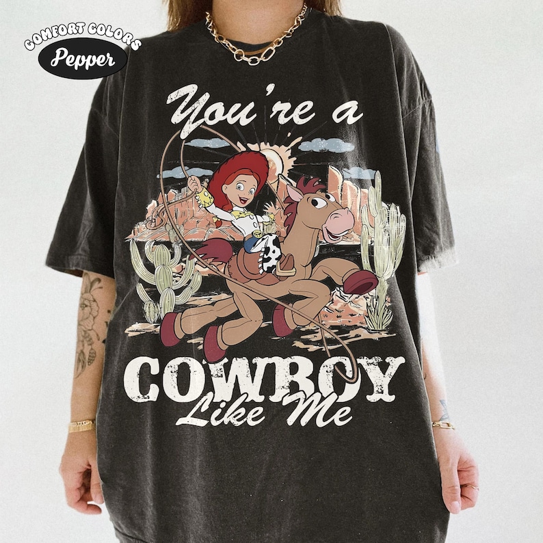 May include: A charcoal black short-sleeve t-shirt featuring a graphic of a cowgirl riding a brown horse with a lasso. The text reads You're a COWBOY Like Me. The design includes desert scenery with cacti and mountains in a distressed, vintage style.