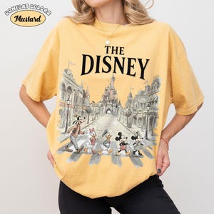 May include: Mustard-colored t-shirt with the text "THE DISNEY" above a graphic of Disney characters crossing a street in front of a castle. The shirt has the Comfort Colors logo.