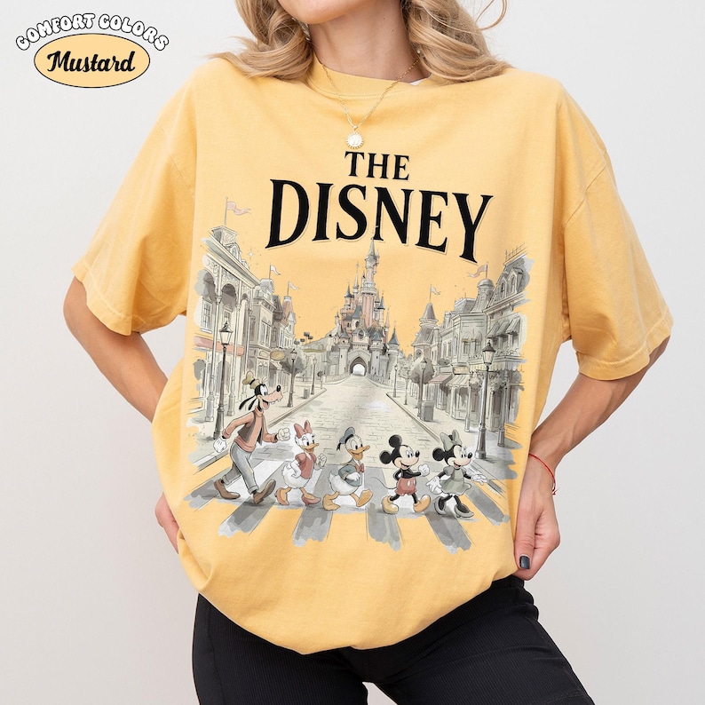 May include: Mustard-colored t-shirt with the text "THE DISNEY" above a graphic of Disney characters crossing a street in front of a castle. The shirt has the Comfort Colors logo.