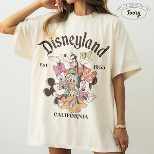 May include: Ivory-colored t-shirt with a Disneyland graphic. The design features Mickey Mouse, Minnie Mouse, Donald Duck, Daisy Duck, Goofy, and Pluto. The text "Disneyland" is arched above the characters, with "Est 1955" below. "California" is printed at the bottom.