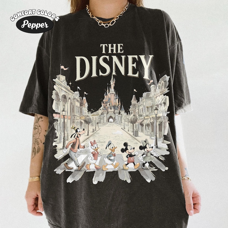 May include: Black Comfort Colors t-shirt featuring a graphic of Disney characters crossing a street in front of a castle. The text "THE DISNEY" is above the image. The shirt has a vintage, distressed look.