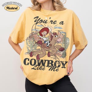 May include: A mustard yellow short-sleeve t-shirt featuring a graphic of the character Jessie riding Bullseye from Toy Story. The design includes a lasso, desert landscape, and the text "You're a COWBOY Like Me" in a distressed black font.
