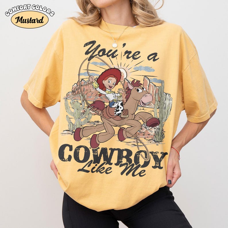 May include: A mustard yellow short-sleeve t-shirt featuring a graphic of the character Jessie riding Bullseye from Toy Story. The design includes a lasso, desert landscape, and the text "You're a COWBOY Like Me" in a distressed black font.
