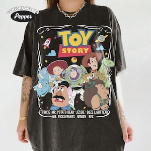 May include: Dark gray Comfort Colors t-shirt featuring the Toy Story logo and character illustrations. The design includes Woody, Buzz Lightyear, Jessie, Mr. Potato Head, Rex, and others. The text at the bottom lists the characters' names.