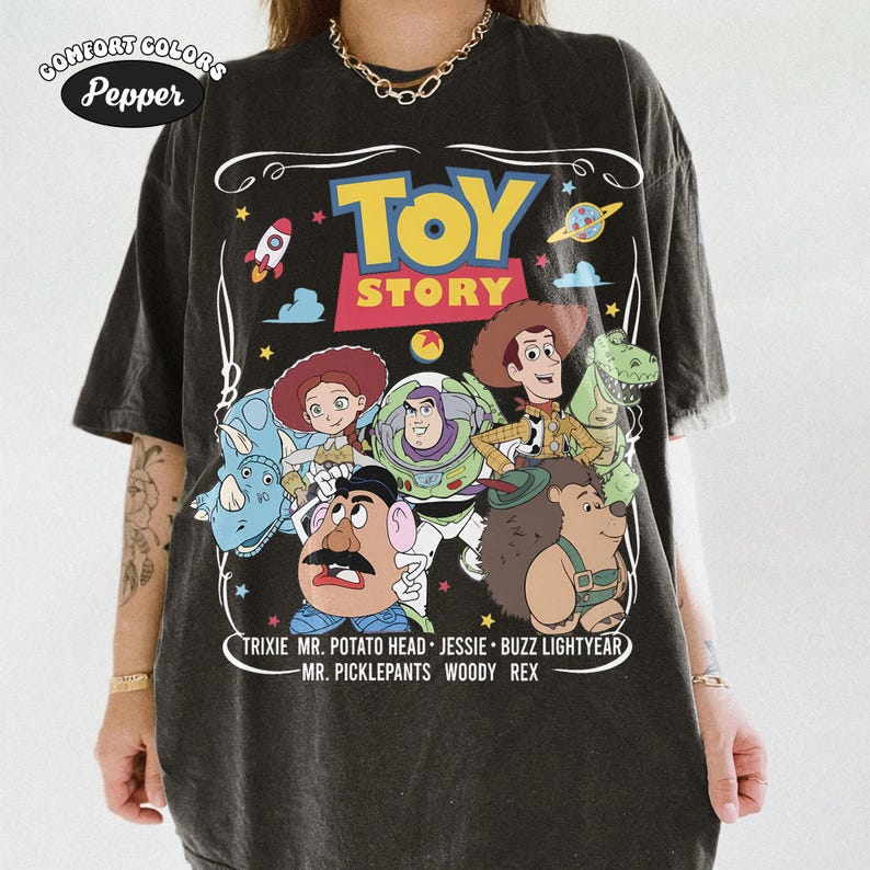 May include: Dark gray Comfort Colors t-shirt featuring the Toy Story logo and character illustrations. The design includes Woody, Buzz Lightyear, Jessie, Mr. Potato Head, Rex, and others. The text at the bottom lists the characters' names.