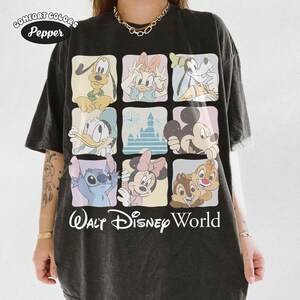 Op de afbeelding: Een antracietzwart t-shirt met een raster van negen vierkanten met illustraties van diverse tekenfilmfiguren en een kasteel. De tekst "Walt Disney World" staat onderaan in het wit gedrukt. Een "Comfort Colors Pepper"-logo staat linksboven.