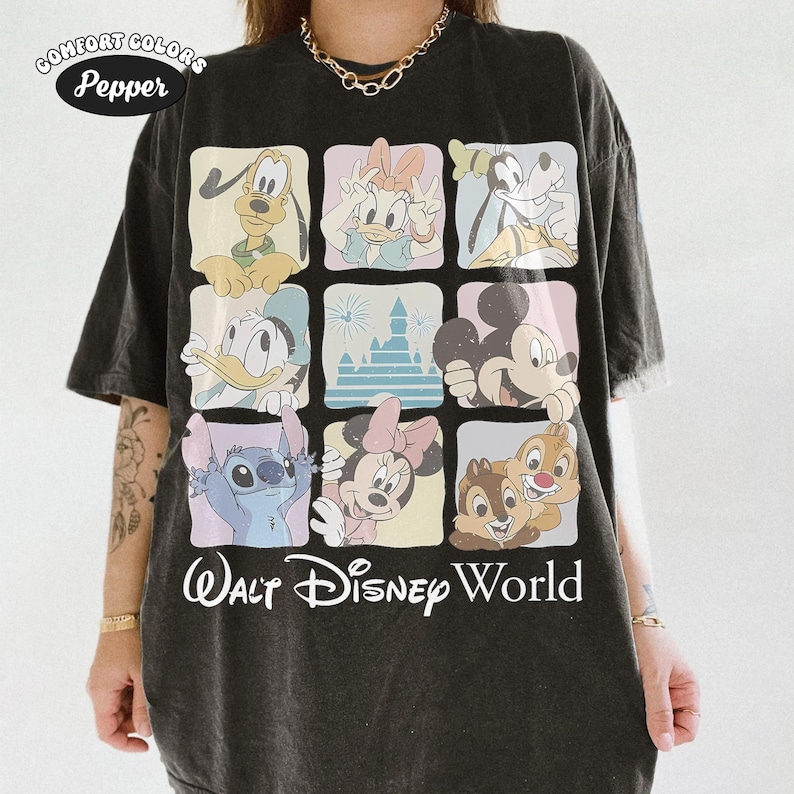 Op de afbeelding: Een antracietzwart t-shirt met een raster van negen vierkanten met illustraties van diverse tekenfilmfiguren en een kasteel. De tekst "Walt Disney World" staat onderaan in het wit gedrukt. Een "Comfort Colors Pepper"-logo staat linksboven.