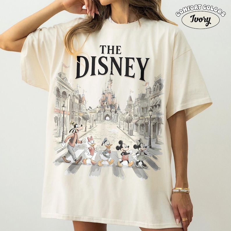 May include: Ivory-colored Comfort Colors t-shirt featuring a graphic of Disney characters crossing a street in front of a castle. The text "THE DISNEY" is printed above the image. The shirt has short sleeves.