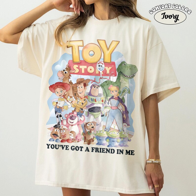May include: Ivory-colored t-shirt featuring a colorful graphic of Toy Story characters. The design includes Woody, Buzz Lightyear, and other characters from the movie, with the text "TOY STORY" and "YOU'VE GOT A FRIEND IN ME".