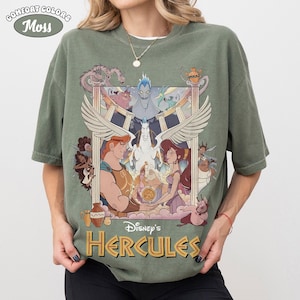 May include: Moss green Comfort Colors t-shirt featuring a colorful graphic from Disney's Hercules. The design includes Hercules, Megara, Pegasus, and Hades. The words "Disney's Hercules" are printed in gold.