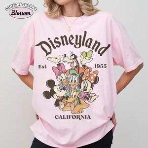 May include: Pink t-shirt with a Disneyland graphic featuring Mickey Mouse, Minnie Mouse, Donald Duck, Daisy Duck, Goofy, and Pluto. The text "Disneyland" arches above the characters, with "Est 1955" and "California" below.