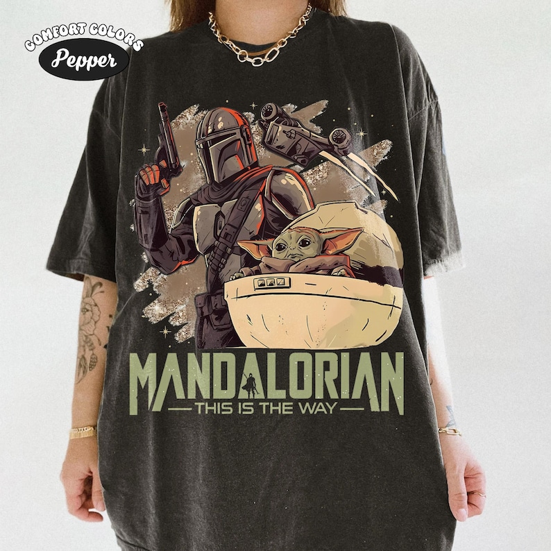 May include: A charcoal gray t-shirt featuring a graphic print of a Mandalorian character holding a blaster, a small green creature in a floating pod, and a spaceship. The text reads MANDALORIAN THIS IS THE WAY. The shirt has a Comfort Colors Pepper label.