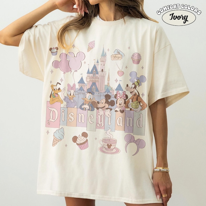 May include: Ivory-colored t-shirt featuring a Disneyland graphic. The design includes a castle, cartoon characters, and the word "Disneyland" in pastel colors. Additional graphics include ice cream, cupcakes, and Mickey Mouse ears.