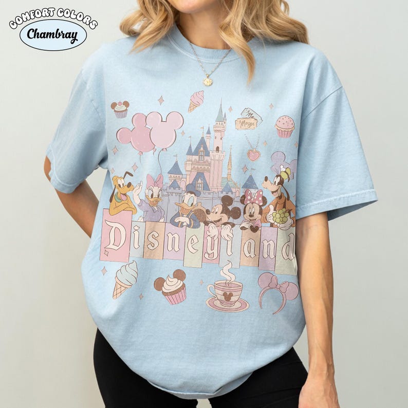 May include: Light blue t-shirt with a Disneyland graphic featuring cartoon characters and a castle. The word "Disneyland" is displayed in a pastel font. The shirt has short sleeves and a crew neck.