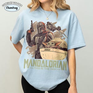 May include: A light blue t-shirt featuring a graphic print of a Mandalorian character holding a blaster, a small green creature in a pod, and a starship. The text "MANDALORIAN" and "THIS IS THE WAY" is printed below the graphic in a distressed font.