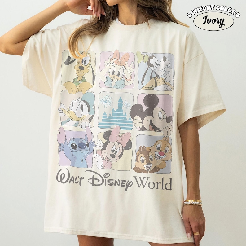 Op de afbeelding: Een ivoorkleurig t-shirt met een raster van negen vierkanten met diverse stripfiguren en een kasteel, met de tekst Walt Disney World eronder gedrukt. De rechterbovenhoek bevat een cirkelvormig logo met de tekst Comfort Colors Ivory.