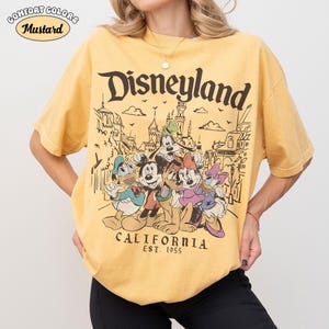 May include: Mustard-colored t-shirt with a Disneyland graphic. The graphic features Mickey Mouse, Donald Duck, Goofy, Minnie Mouse, and Daisy Duck in front of a Disneyland scene. The text "Disneyland California Est. 1955" is also printed on the shirt.