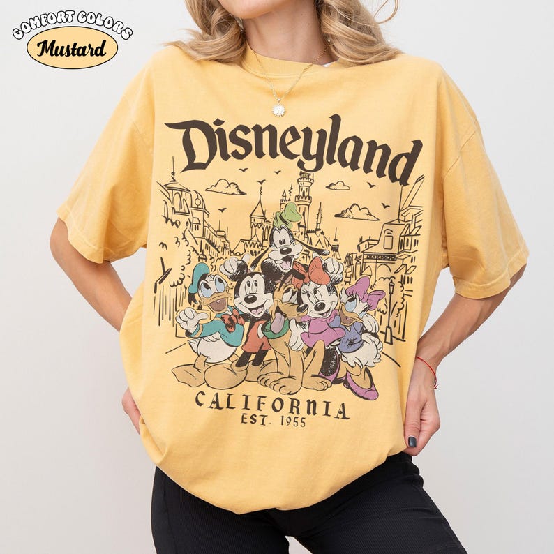 May include: Mustard-colored t-shirt with a Disneyland graphic. The graphic features Mickey Mouse, Donald Duck, Goofy, Minnie Mouse, and Daisy Duck in front of a Disneyland scene. The text "Disneyland California Est. 1955" is also printed on the shirt.