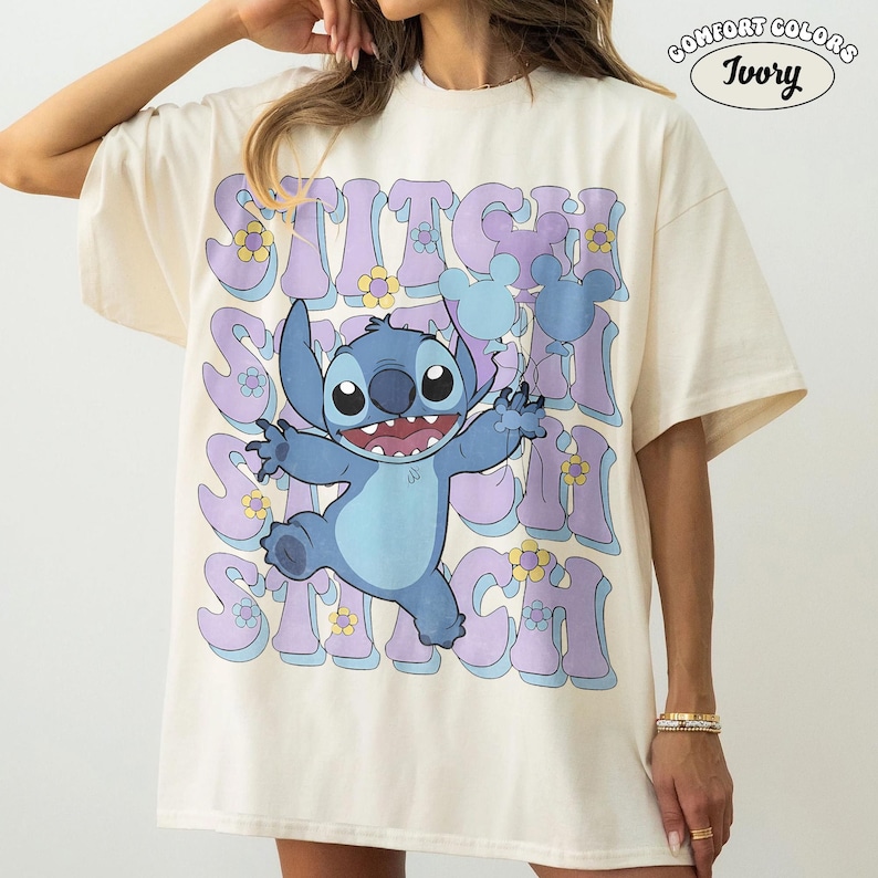 May include: An ivory t-shirt featuring a large graphic of the blue character Stitch from Lilo & Stitch. The design includes repeating purple text, small yellow flowers, and light blue Mickey Mouse-shaped balloons. The text Comfort Colors Ivory is visible.
