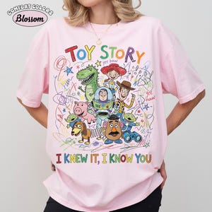 Puede incluir: Una camiseta rosa claro con un gr&aacute;fico colorido de los personajes de Toy Story, incluyendo a Woody, Buzz Lightyear, Rex, Jessie, Hamm, Mr. Potato Head, Slinky Dog y los Aliens. El dise&ntilde;o incluye garabatos, estrellas y el texto TOY STORY e I KNEW IT, I KNOW YOU.