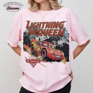 May include: A light pink t-shirt featuring a graphic print of the character Lightning McQueen from the movie Cars. The design includes the text Lightning McQueen in large letters, the number 95 on the car, and the Disney Pixar Cars logo below.