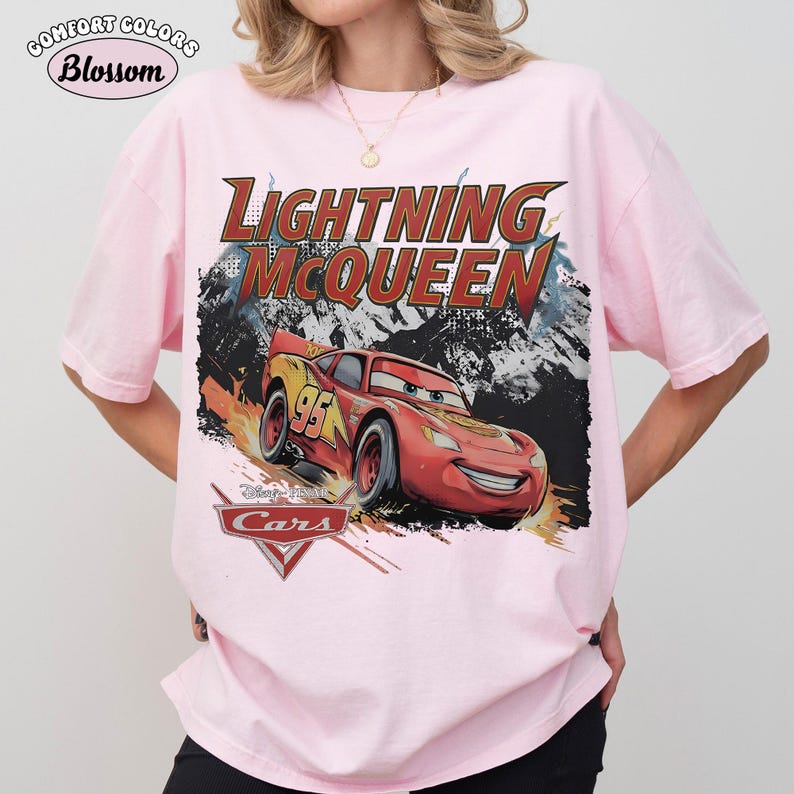 May include: A light pink t-shirt featuring a graphic print of the character Lightning McQueen from the movie Cars. The design includes the text Lightning McQueen in large letters, the number 95 on the car, and the Disney Pixar Cars logo below.
