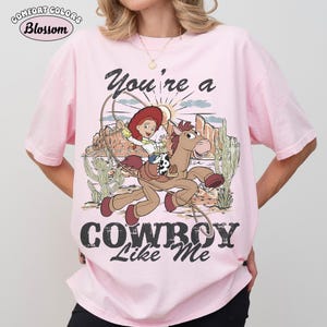 May include: A light pink t-shirt featuring a graphic print of a cowgirl riding a brown horse in a desert landscape with cacti. The text reads "You're a COWBOY Like Me" in black lettering. The design includes a lasso and a sunburst background.