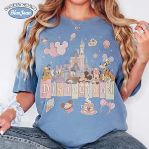 May include: Blue Comfort Colors t-shirt with a Disneyland graphic. The design features the Disneyland castle, Mickey and Minnie Mouse, Donald and Daisy Duck, Goofy, and other Disney icons. The word "Disneyland" is printed across the bottom.