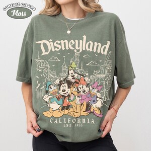 May include: A moss green t-shirt featuring the word "Disneyland" above a graphic of Disney characters and park buildings. The graphic includes Mickey Mouse, Donald Duck, Goofy, and others. The text "CALIFORNIA EST. 1955" is printed below the characters.