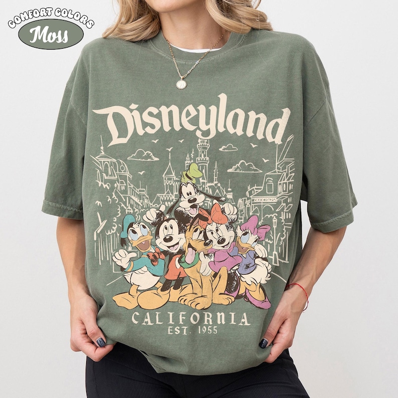 May include: A moss green t-shirt featuring the word "Disneyland" above a graphic of Disney characters and park buildings. The graphic includes Mickey Mouse, Donald Duck, Goofy, and others. The text "CALIFORNIA EST. 1955" is printed below the characters.