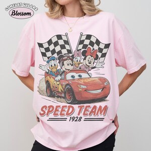 May include: Pale pink Comfort Colors t-shirt with a graphic of Disney characters in a race car and checkered flags. The design includes Mickey Mouse, Donald Duck, Daisy Duck, and Minnie Mouse. The text "SPEED TEAM 1928" is printed below the car.