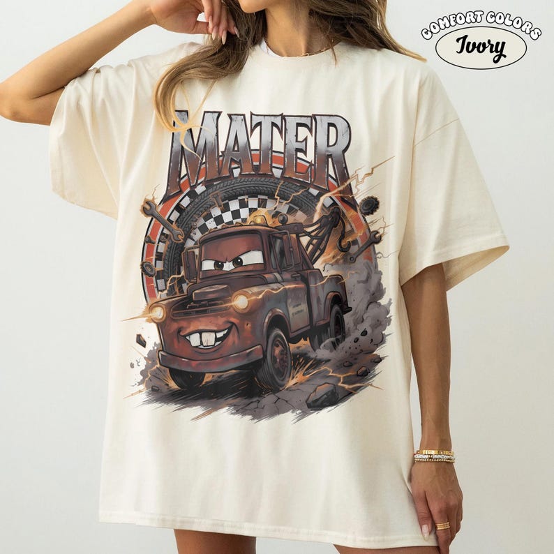 May include: Ivory-colored t-shirt featuring a graphic of Mater from the Cars movie. The design includes the word "MATER" in a distressed font, a checkered flag, and a cartoon image of Mater. The shirt is a Comfort Colors brand.