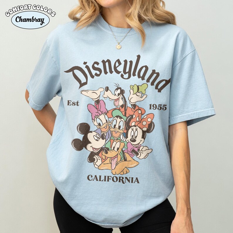 May include: Light blue t-shirt with a vintage Disneyland graphic featuring Mickey Mouse, Minnie Mouse, Donald Duck, Daisy Duck, Goofy, and Pluto. The shirt includes the text "Disneyland California Est 1955" in a brown font. The shirt is made of Comfort Colors Chambray fabric.
