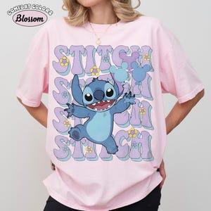 May include: A light pink short-sleeve t-shirt featuring a graphic of the blue character Stitch holding light blue balloons. The background includes the repeated text STITCH in large purple letters with yellow flowers and light blue accents.