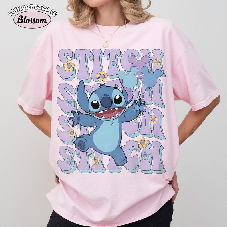 May include: A light pink short-sleeve t-shirt featuring a graphic of the blue character Stitch holding light blue balloons. The background includes the repeated text STITCH in large purple letters with yellow flowers and light blue accents.