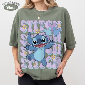 May include: A moss green short-sleeve t-shirt featuring a graphic of the character Stitch holding blue and purple balloons. The background text reads STITCH in large, wavy, light purple letters, accented with small yellow and blue flowers.