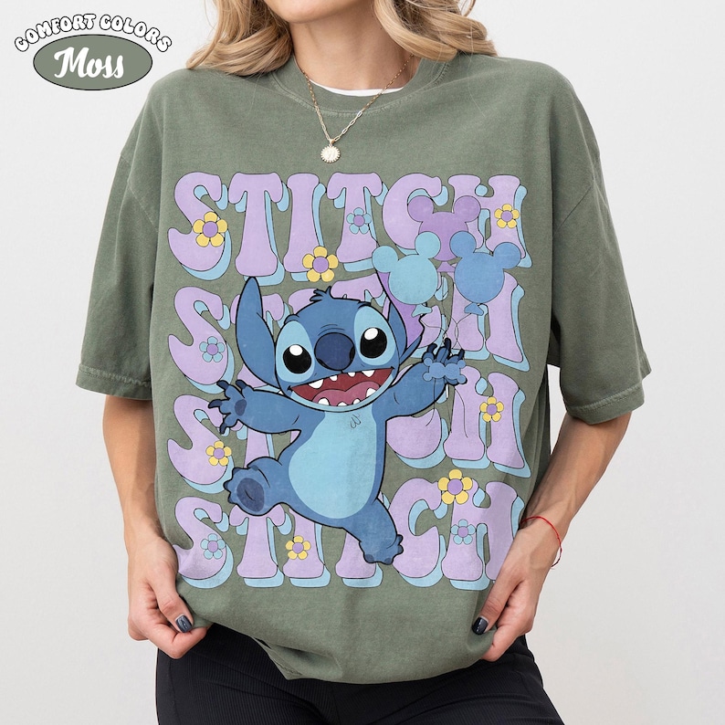 May include: A moss green short-sleeve t-shirt featuring a graphic of the character Stitch holding blue and purple balloons. The background text reads STITCH in large, wavy, light purple letters, accented with small yellow and blue flowers.