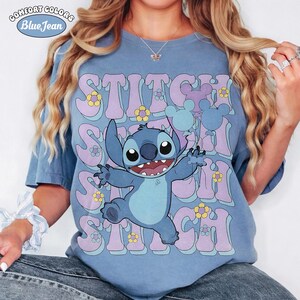 May include: A blue t-shirt featuring a graphic of the character Stitch with his arms outstretched. The background includes repeating purple text that reads STITCH, interspersed with small yellow and white flowers and light blue Mickey Mouse balloons.