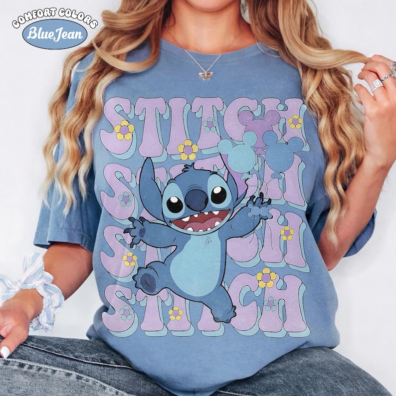 May include: A blue t-shirt featuring a graphic of the character Stitch with his arms outstretched. The background includes repeating purple text that reads STITCH, interspersed with small yellow and white flowers and light blue Mickey Mouse balloons.