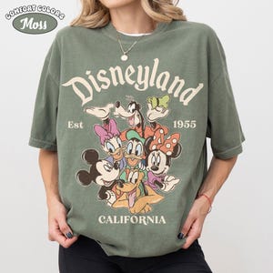 May include: Green Comfort Colors t-shirt with a Disneyland graphic. The graphic features Mickey Mouse, Minnie Mouse, Donald Duck, Daisy Duck, Goofy, and Pluto. The text "Disneyland California Est 1955" is also printed on the shirt.