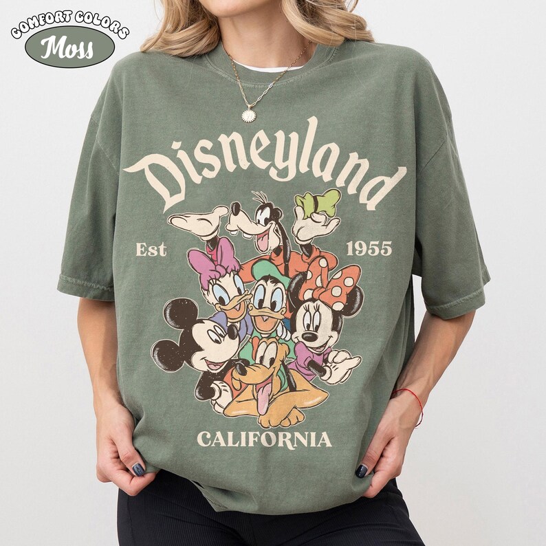 May include: Green Comfort Colors t-shirt with a Disneyland graphic. The graphic features Mickey Mouse, Minnie Mouse, Donald Duck, Daisy Duck, Goofy, and Pluto. The text "Disneyland California Est 1955" is also printed on the shirt.