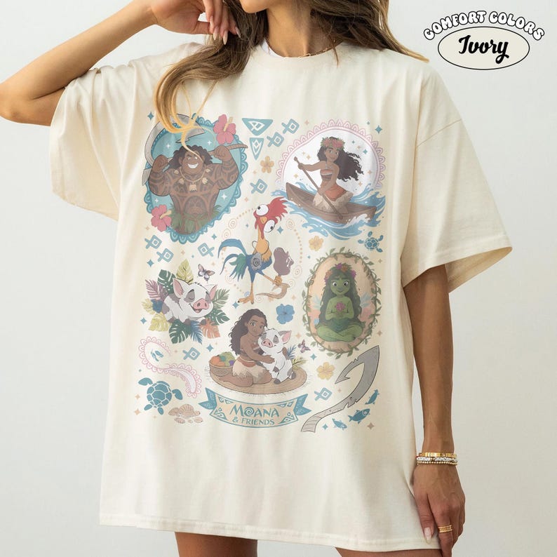 May include: Ivory Comfort Colors t-shirt featuring a colorful graphic of characters from the animated film Moana. The design includes illustrations of Moana, Maui, Pua, Hei Hei, and other elements from the movie. The text "MOANA & FRIENDS" is also included.