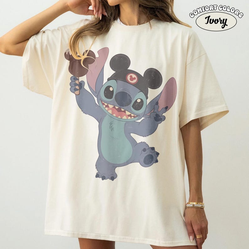 May include: An ivory t-shirt featuring a graphic of the blue character Stitch wearing black mouse ears and holding a brown ice cream bar. The text COMFORT COLORS IVORY is printed in the top right corner. The shirt has short sleeves and a crew neck.
